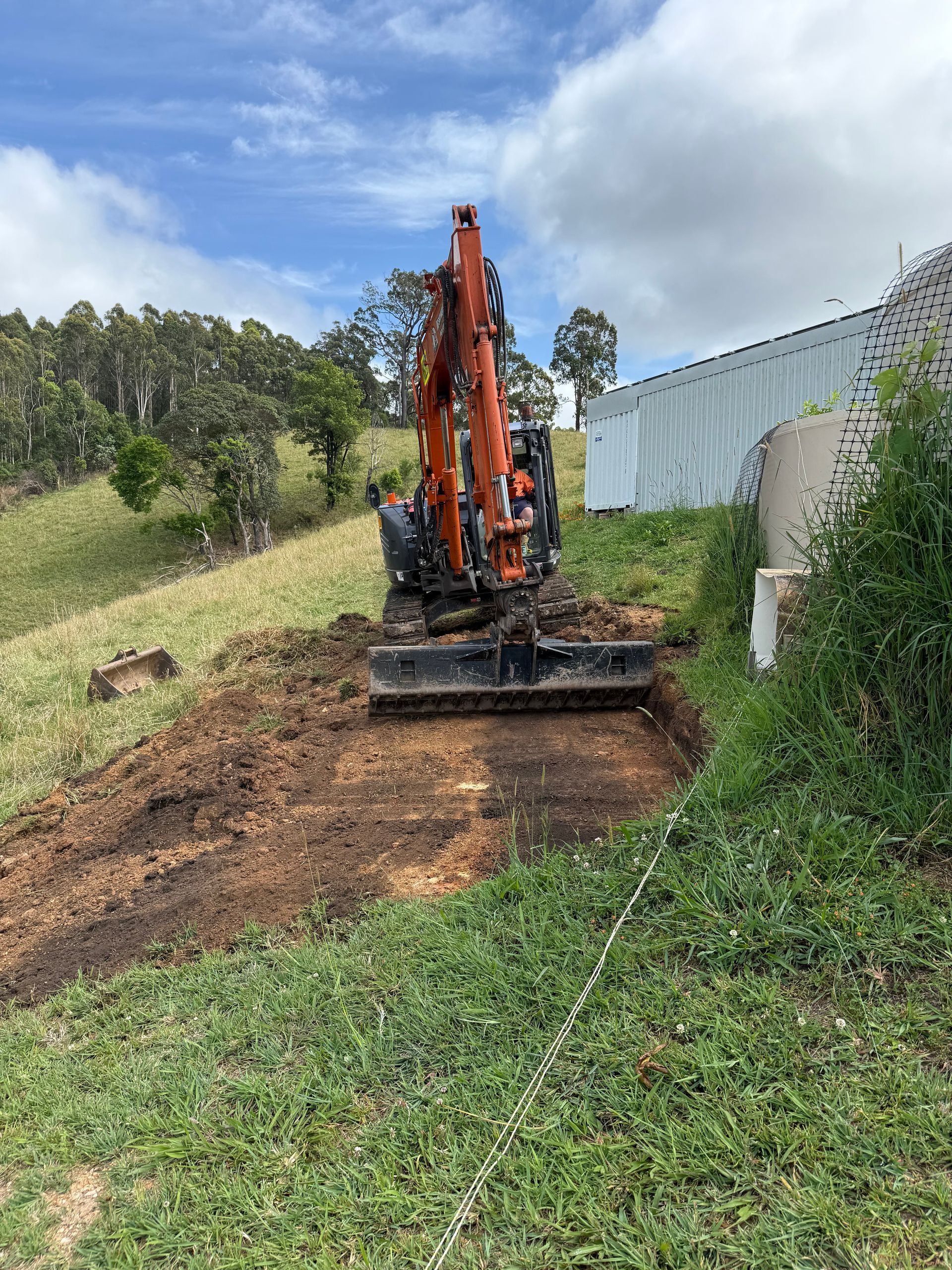 An orange excavator digs into a hillside near a building and water tank.