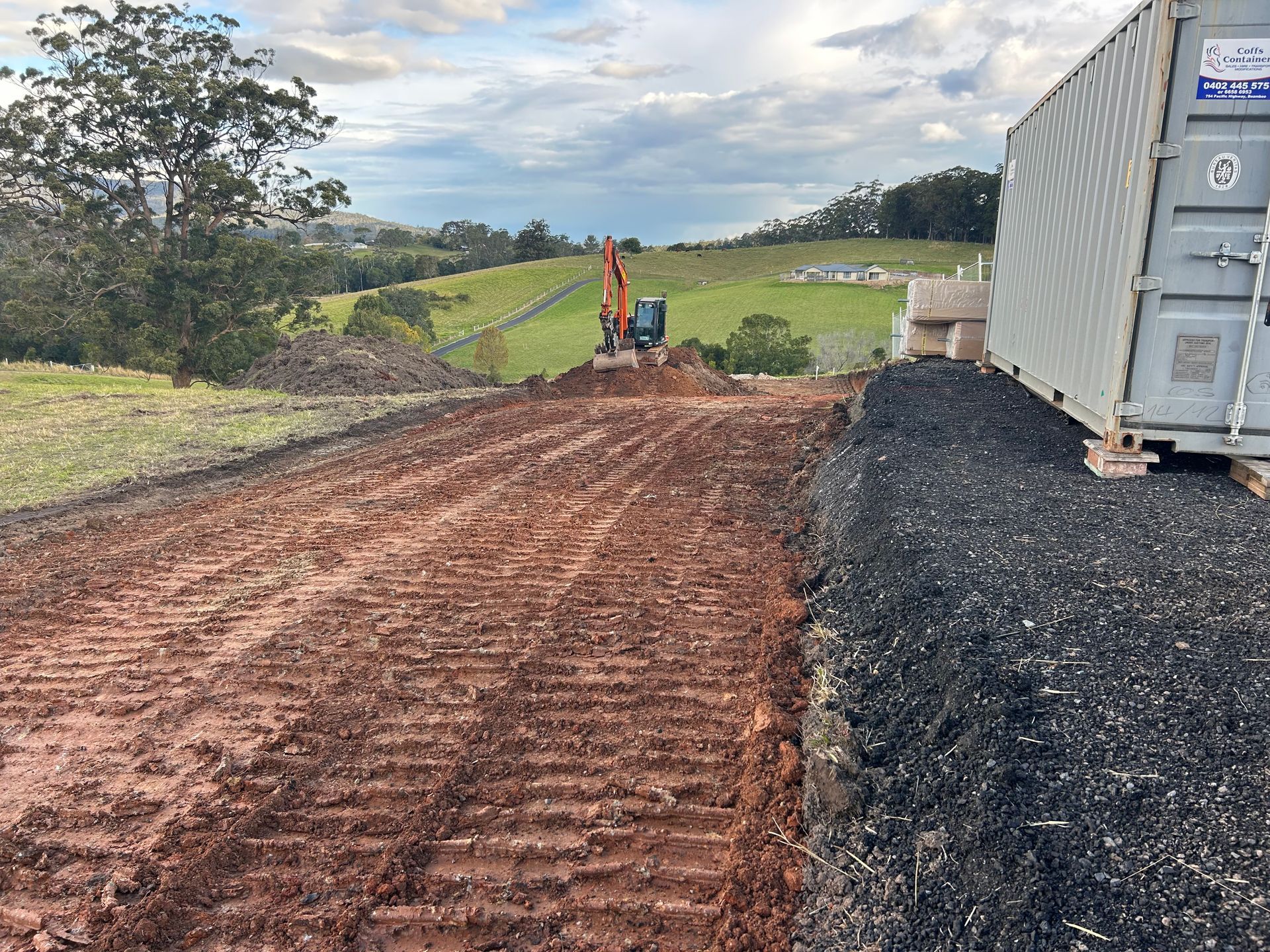 Dirt path with excavator, next to container and green fields under cloudy sky.