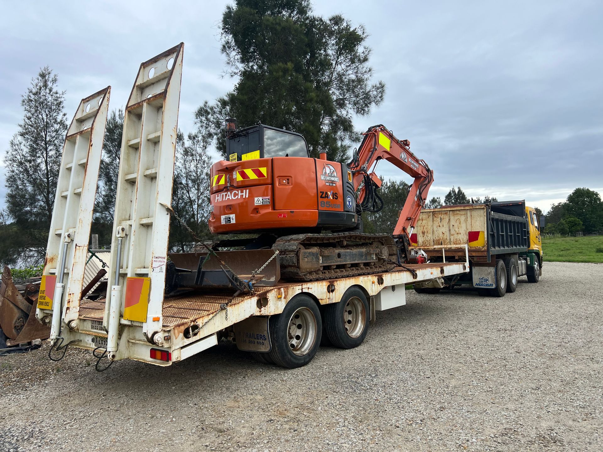 Orange excavator on a flatbed trailer pulled by a yellow truck on a gravel lot under a cloudy sky.