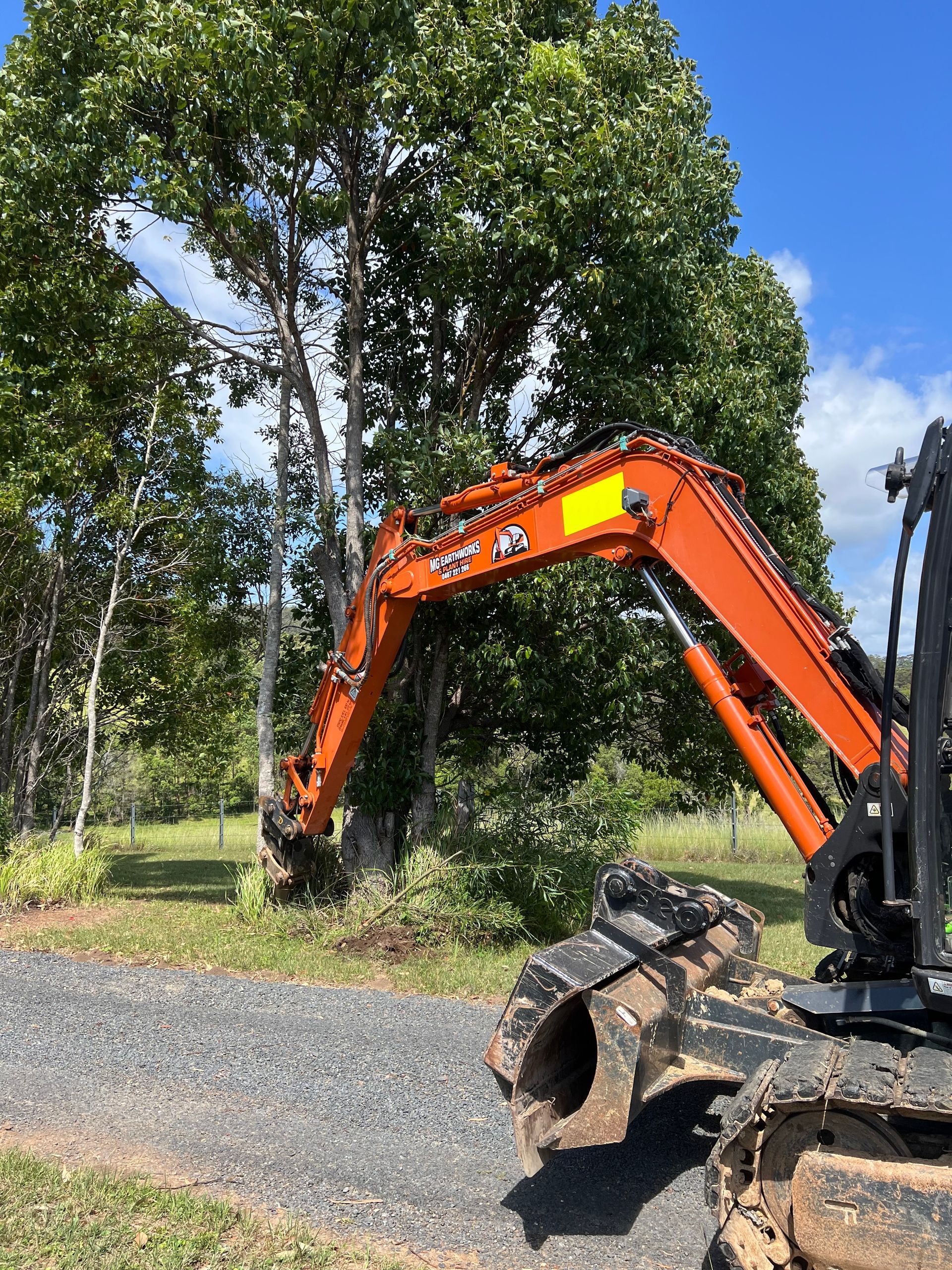 Orange excavator removing a tree; set on a gravel path near a grassy field and green trees under a blue sky.