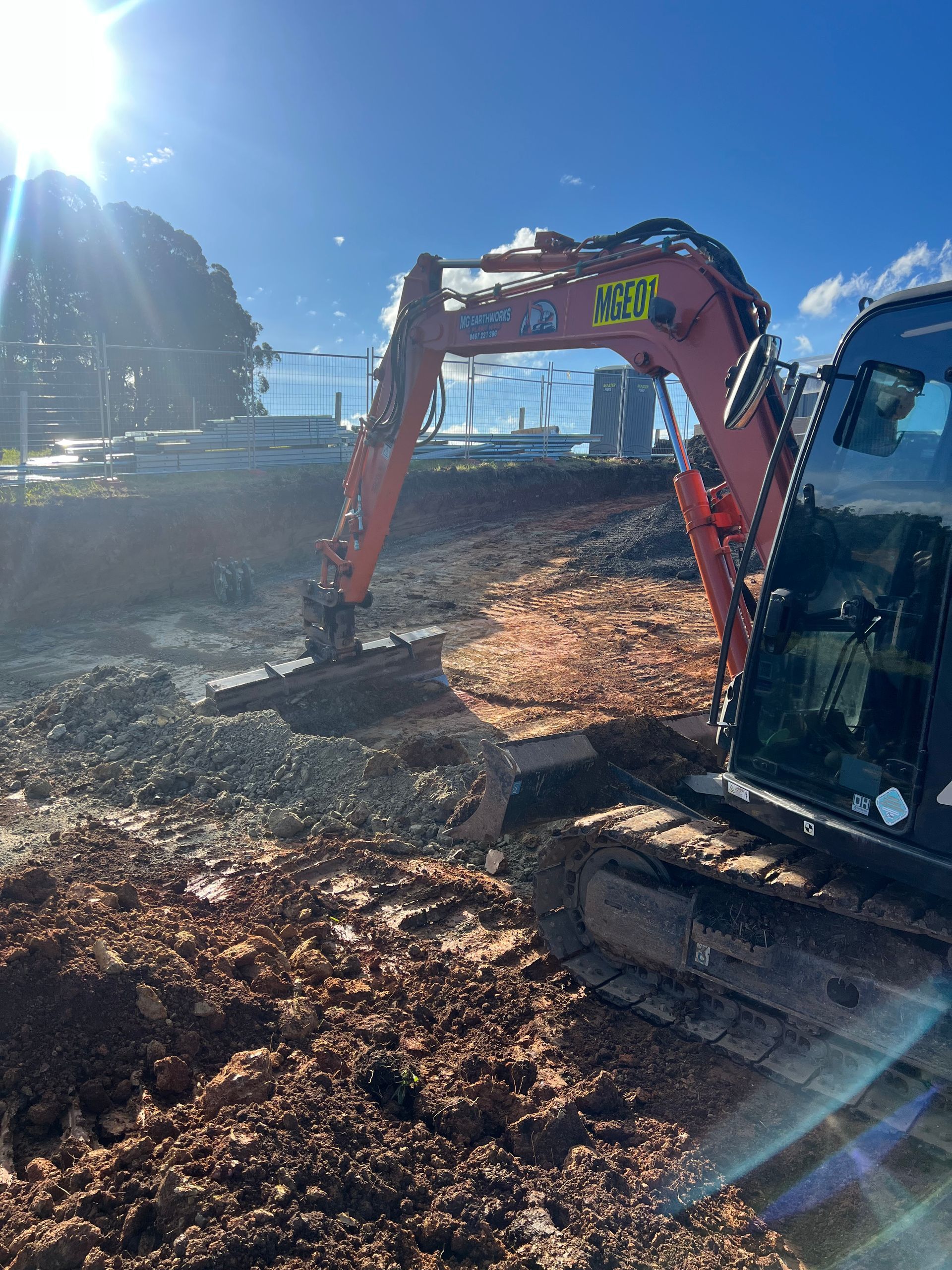 A red excavator digging in a dirt and rock construction site under a bright sun.