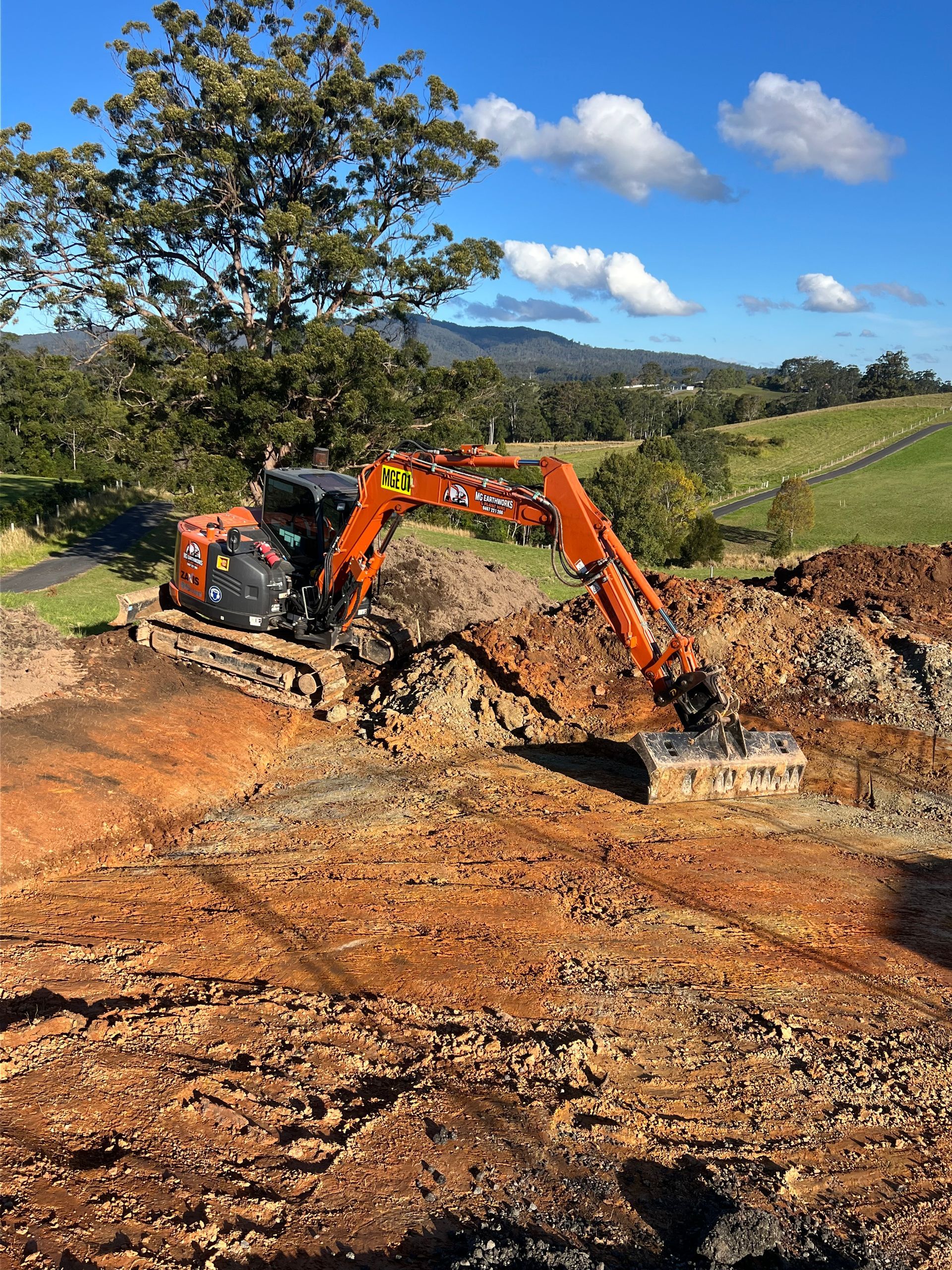 Orange excavator digging into brown earth on a hillside under a cloudy sky.