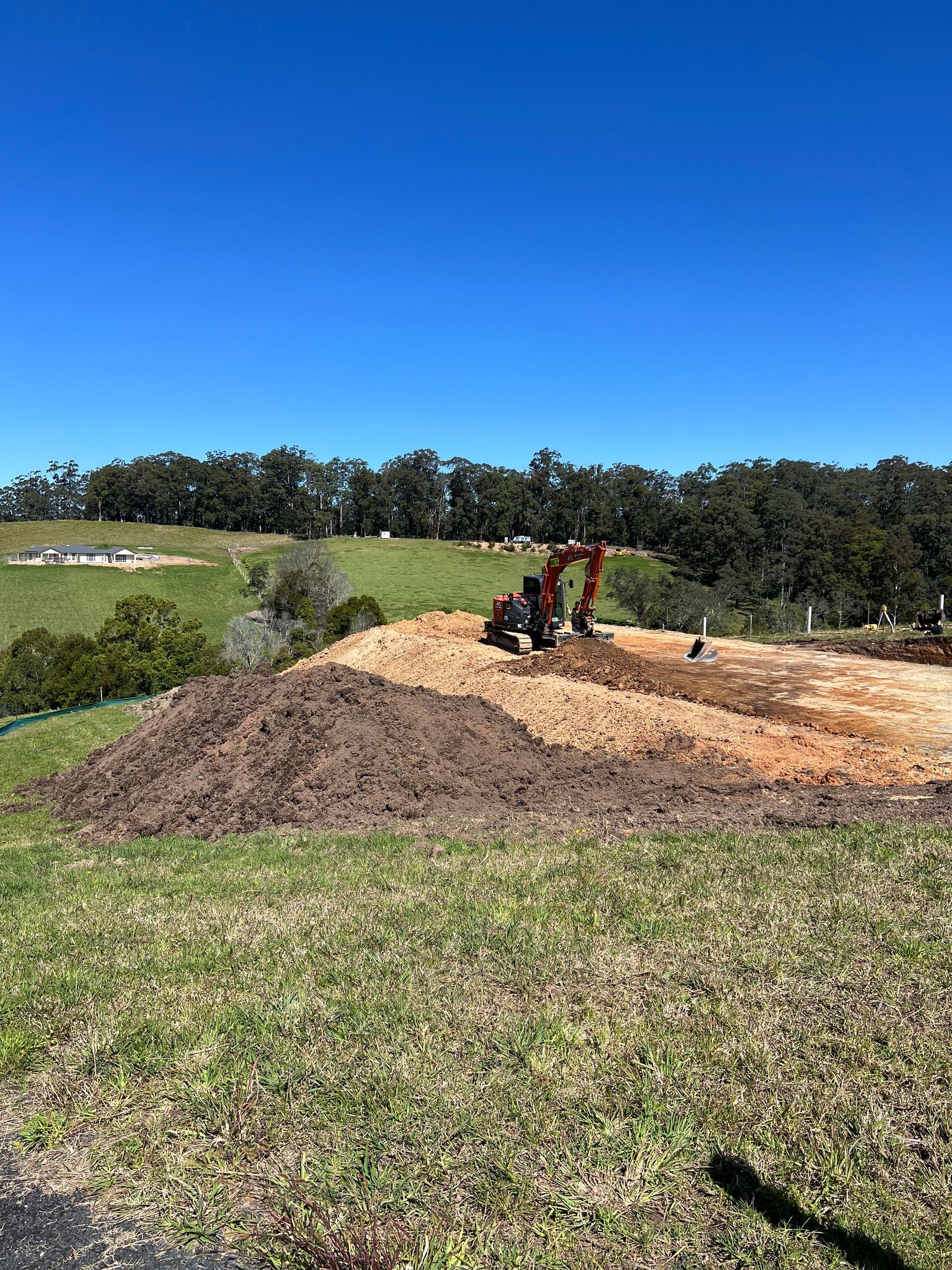 Excavator on a construction site with piles of soil against a backdrop of green hills and a blue sky.