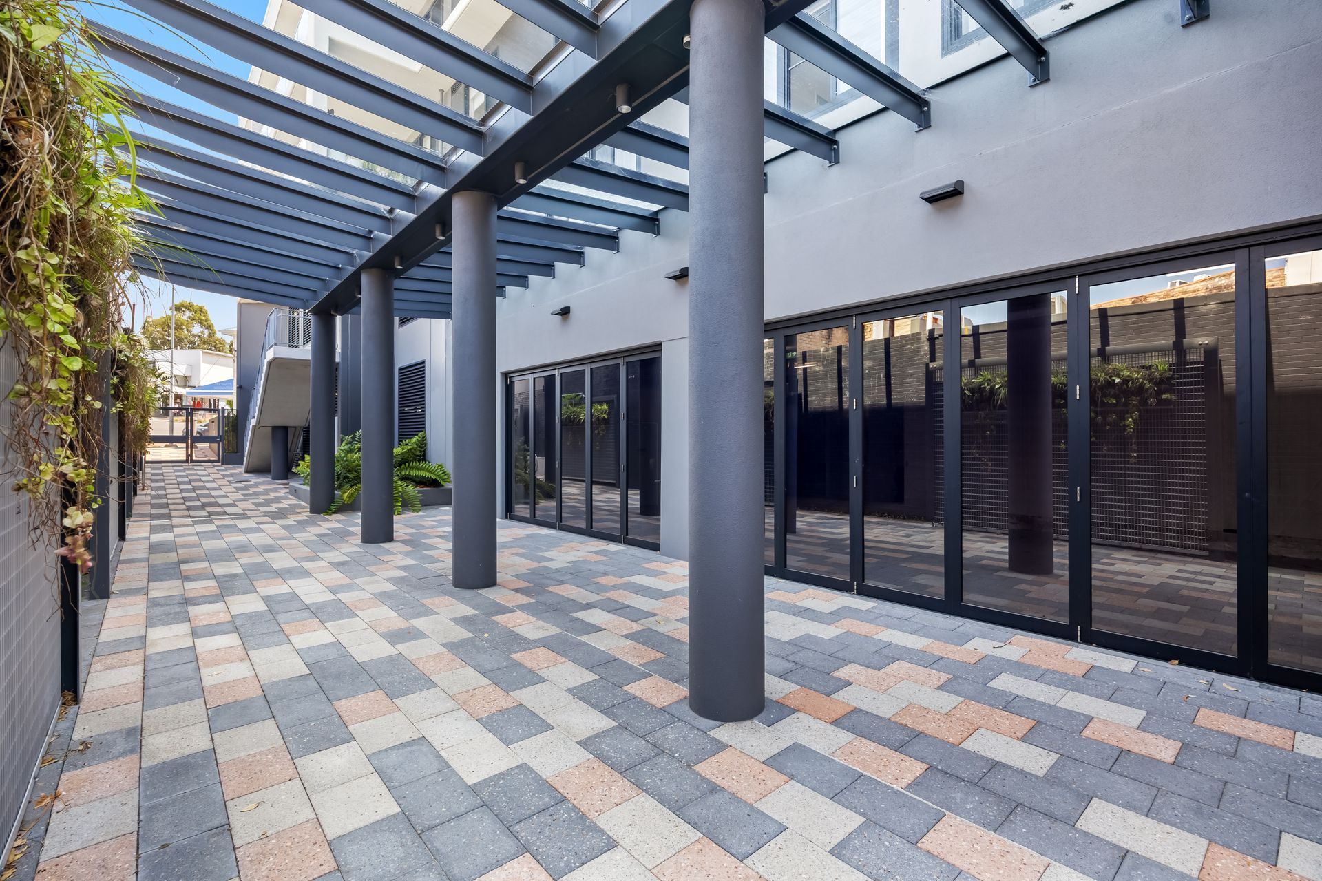 Modern covered outdoor walkway with glass doors, dark columns, and patterned stone paving