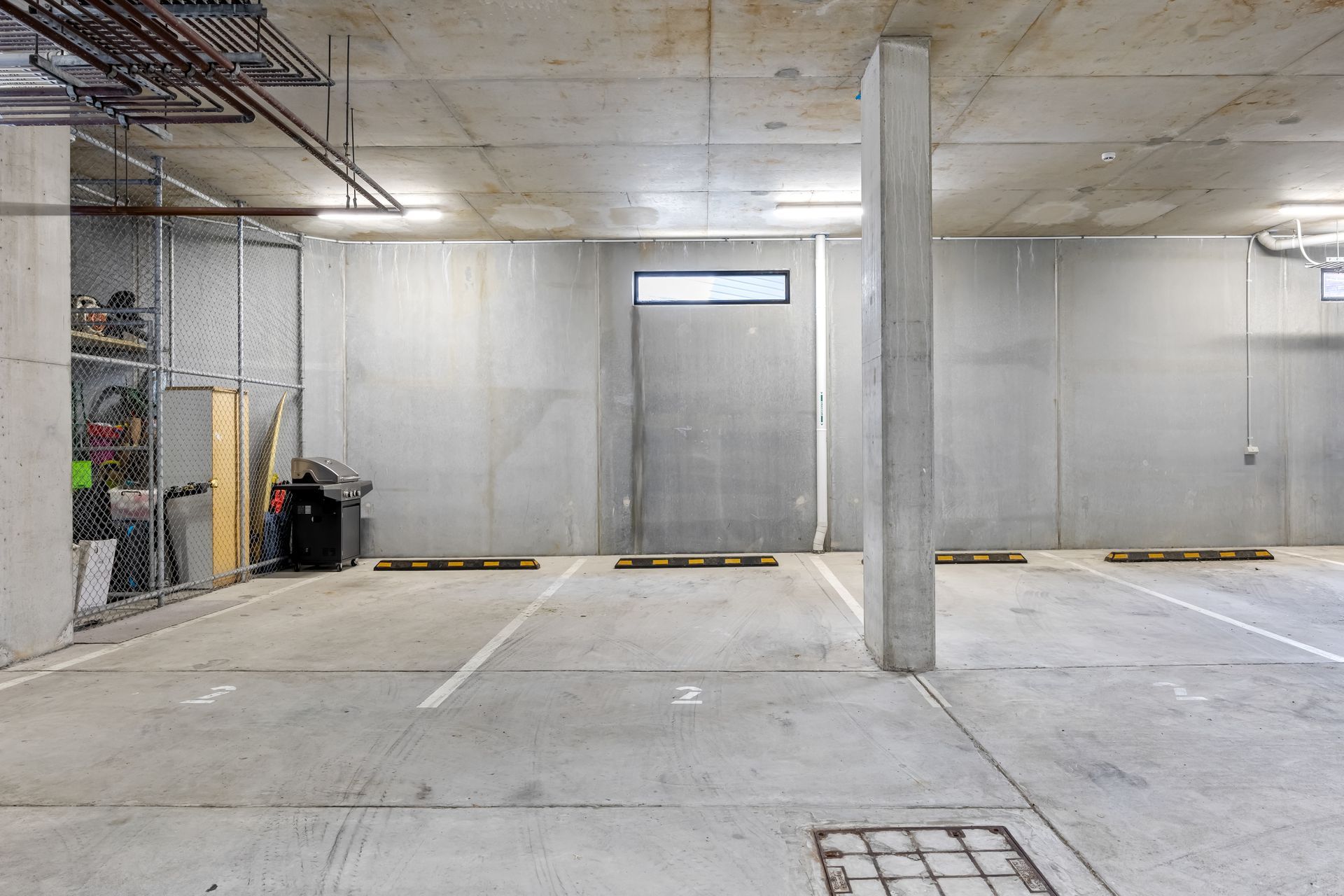 Concrete parking garage with empty spaces, gray walls, a central support column, and a chain-link storage area.