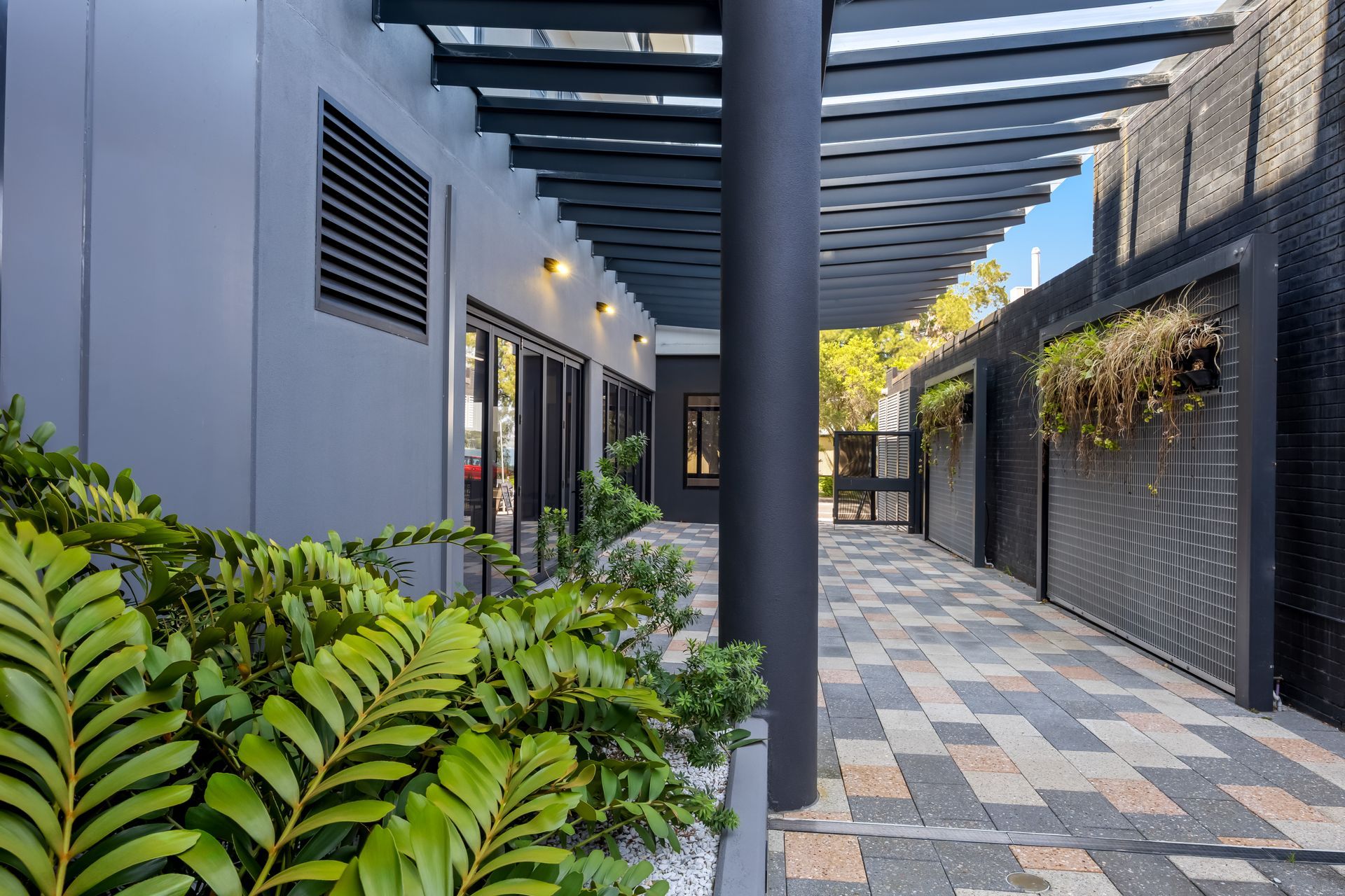 Modern outdoor corridor with gray walls, pavers, pergola beams, and green plants lining the left side
