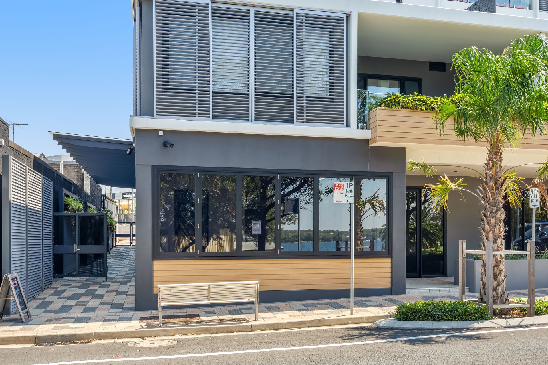 Modern storefront with dark windows, gray facade, wood accents, and a bench on a sunny street