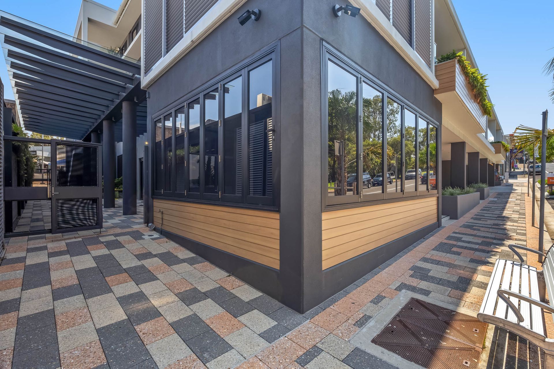 Modern building exterior with large windows, patterned patio paving, awning, and bench-lined walkway