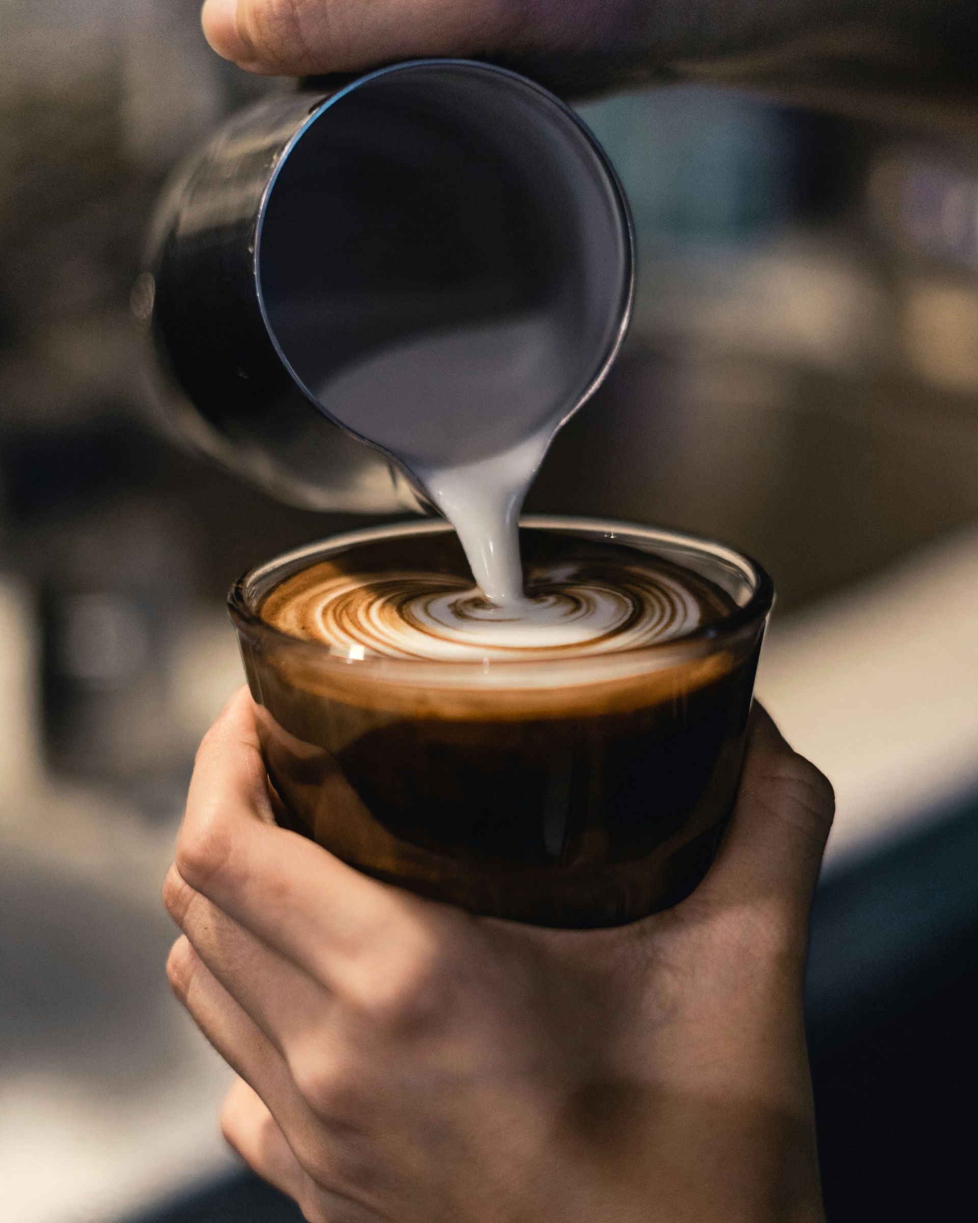 Hand pouring milk into a latte in a dark ceramic cup, creating a swirl pattern