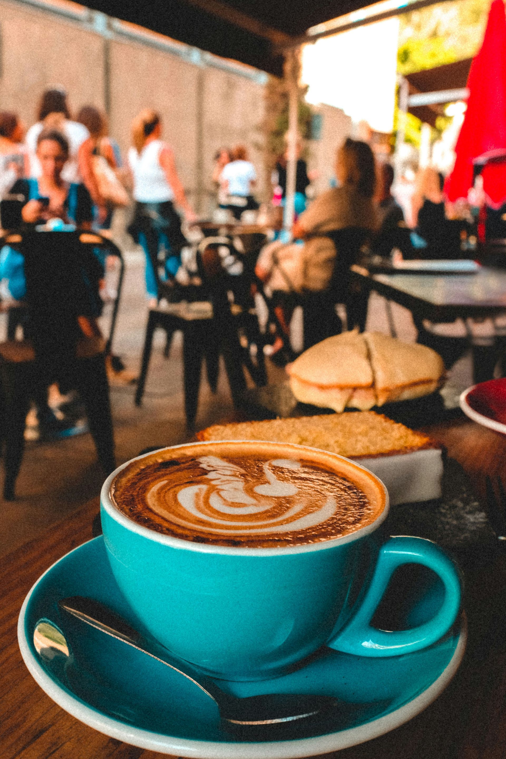 Turquoise cup of latte on a saucer at a café table, with people blurred in the background