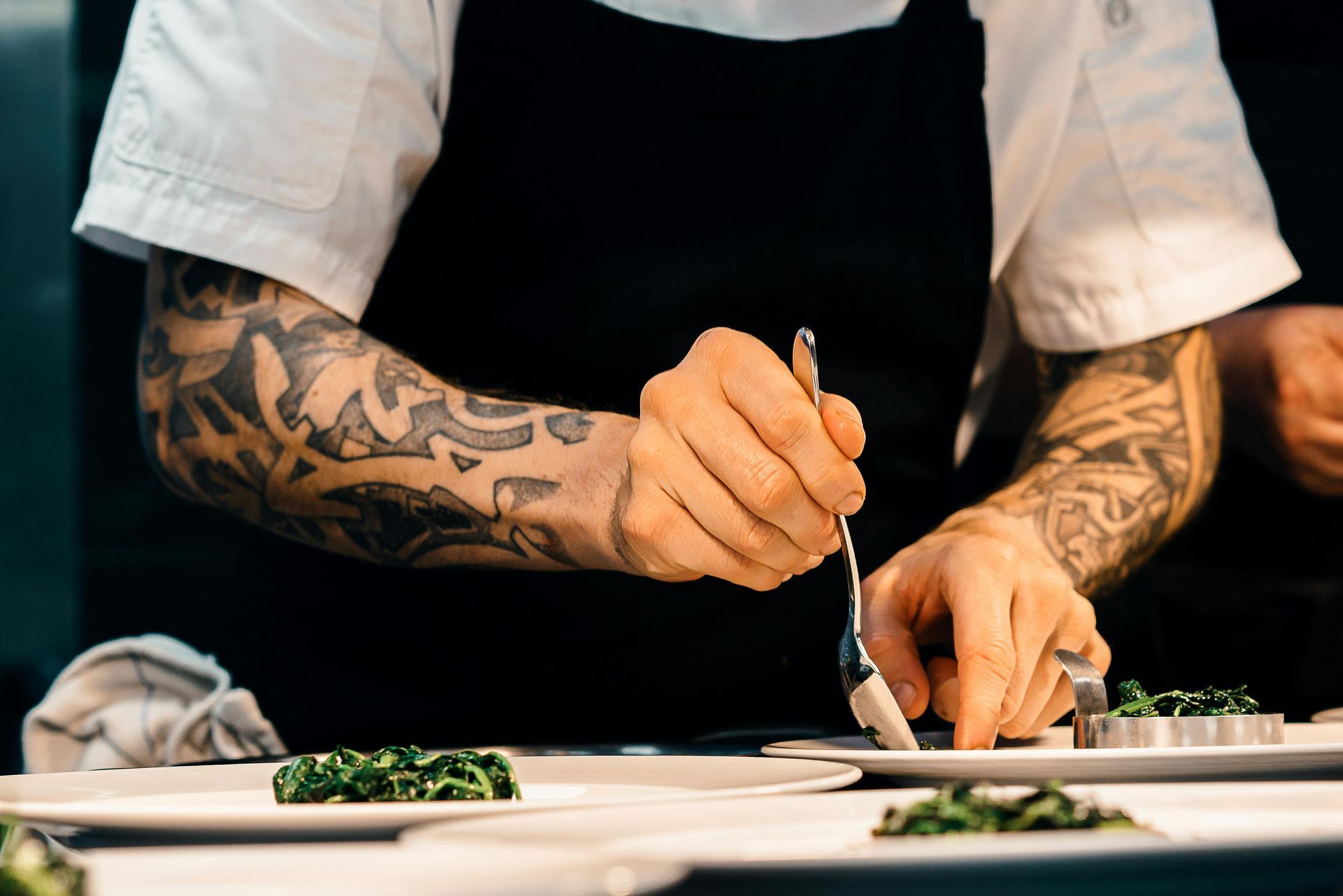 Chef with tattooed arms plating food in a kitchen, using tweezers to arrange herbs on a dish