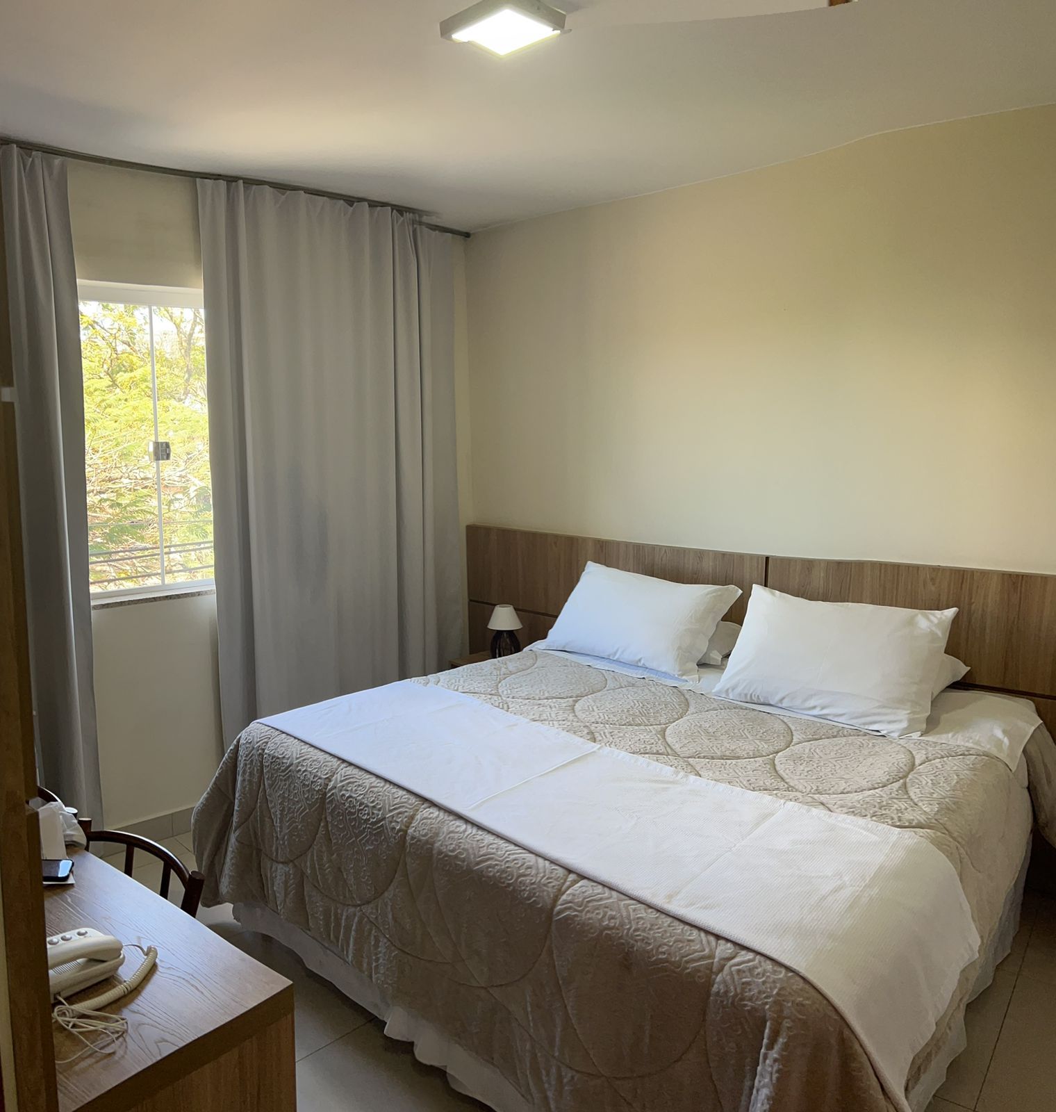 Cama arrumada num quarto luminoso com cortinas cinzentas e vista para a janela.
