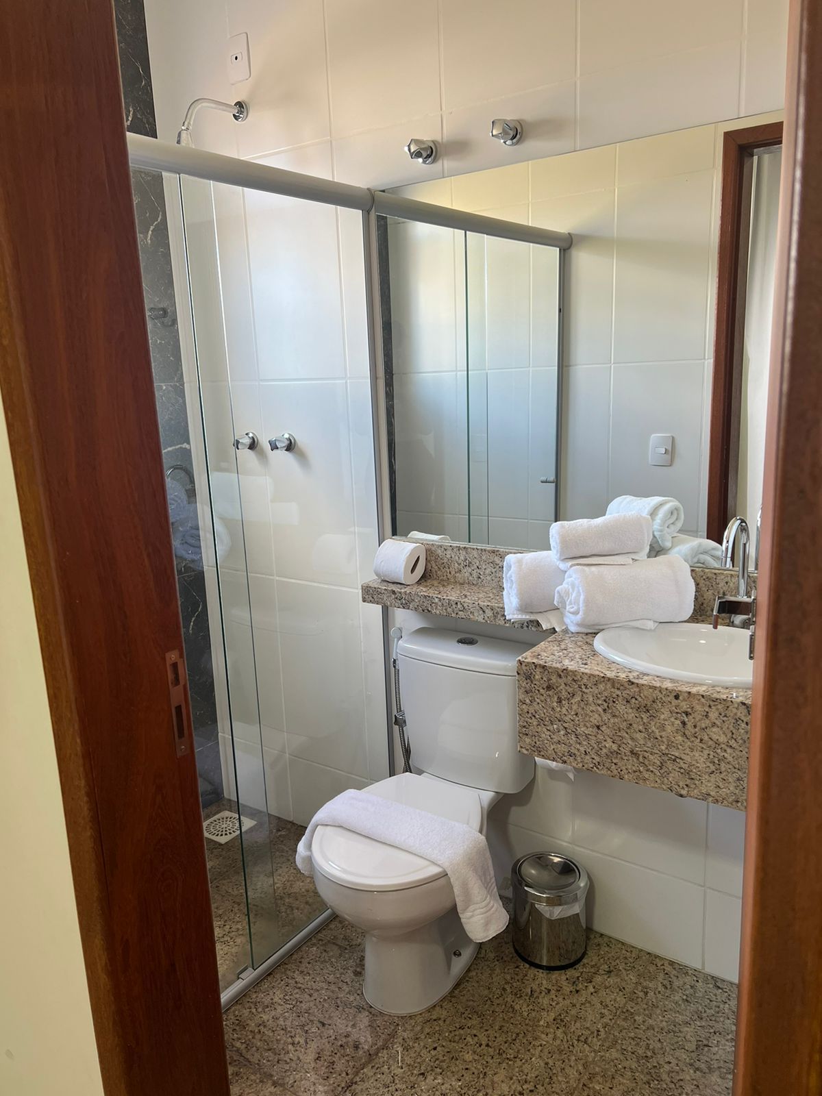 Banheiro pequeno com box de vidro, vaso sanitário e pia de granito com toalhas dobradas.