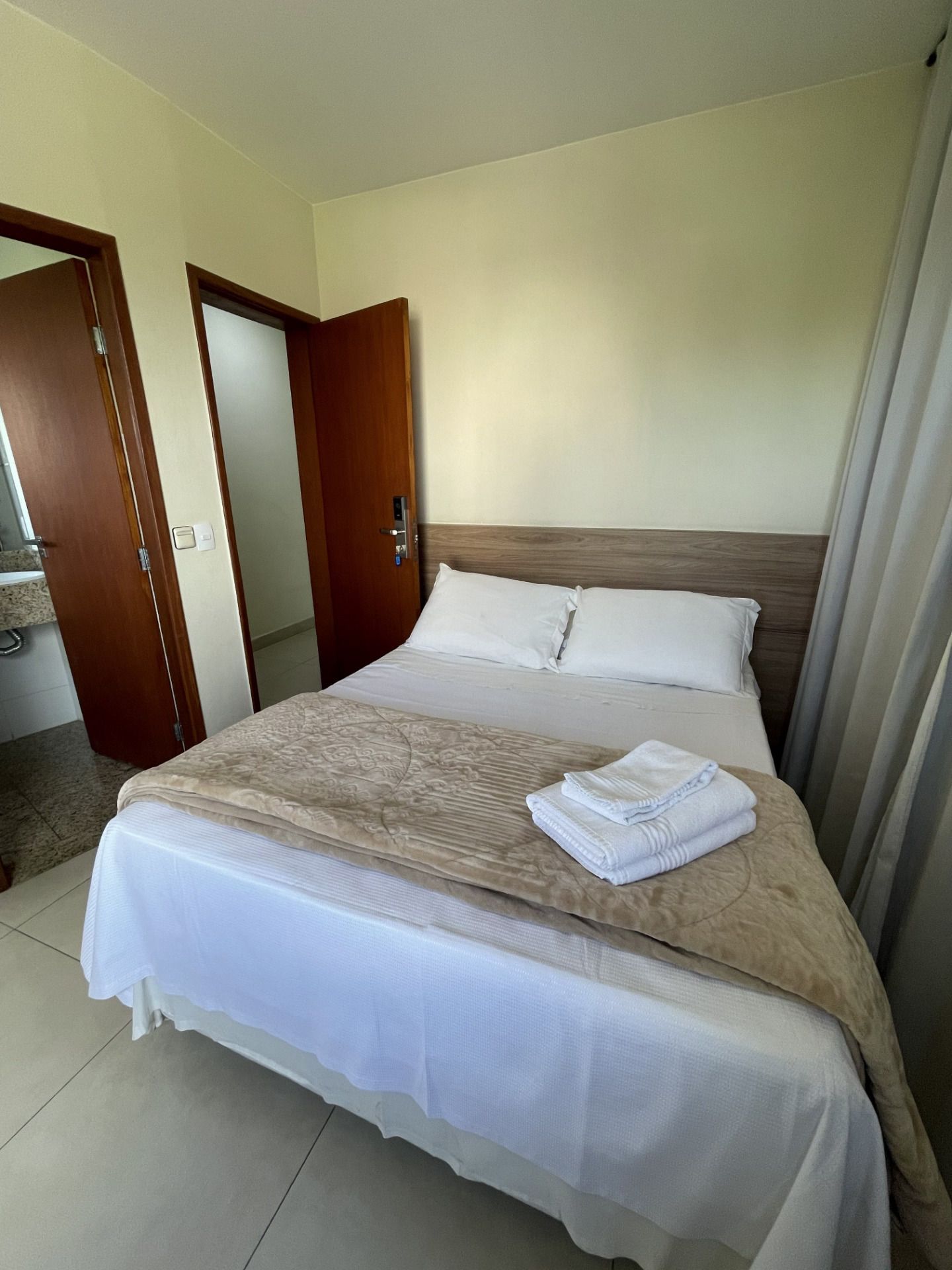Quarto pequeno com cama arrumada, cobertor bege e portas de madeira abertas.