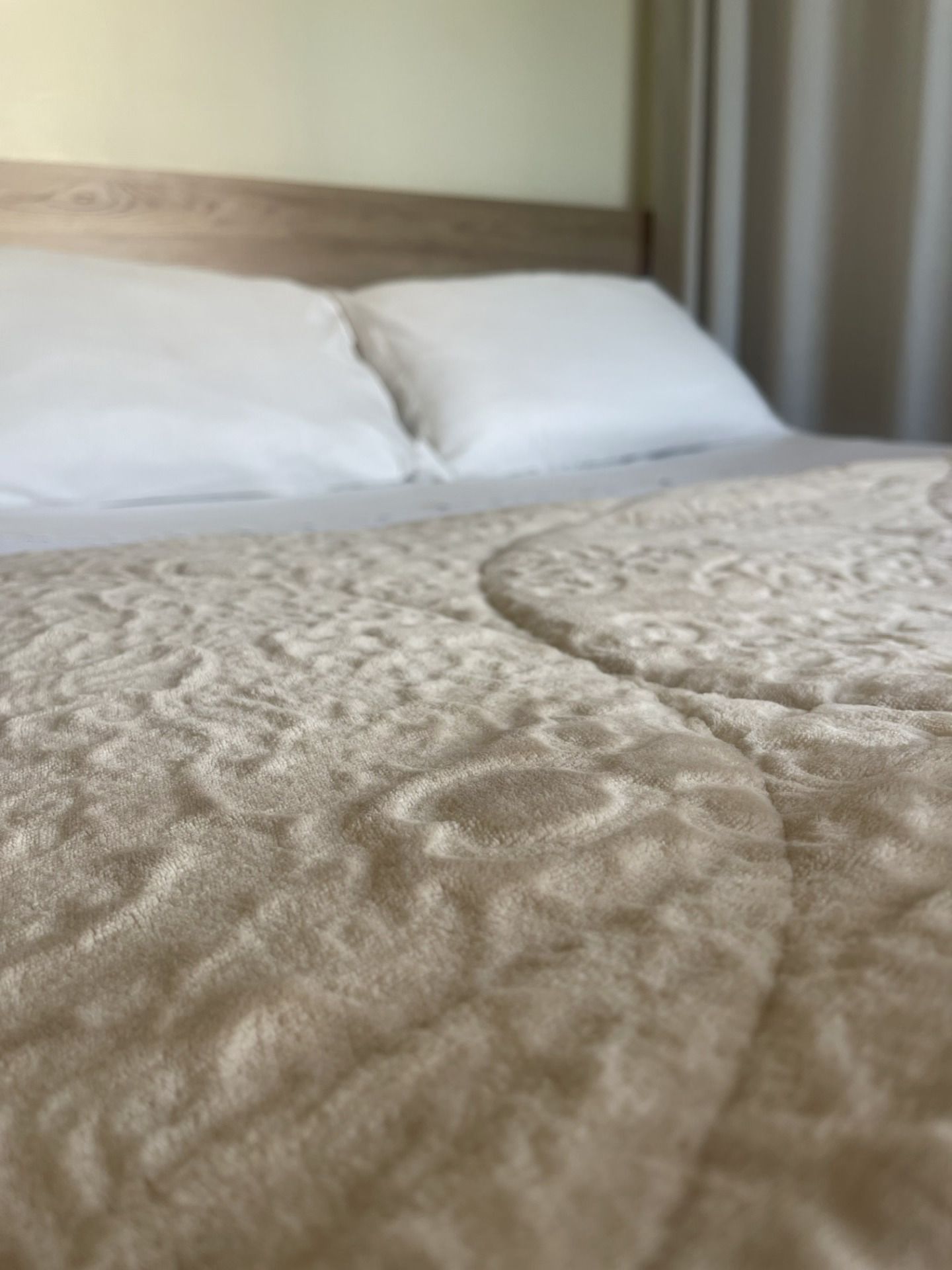 Cama arrumada com travesseiros brancos e colcha bege em um quarto com iluminação suave.