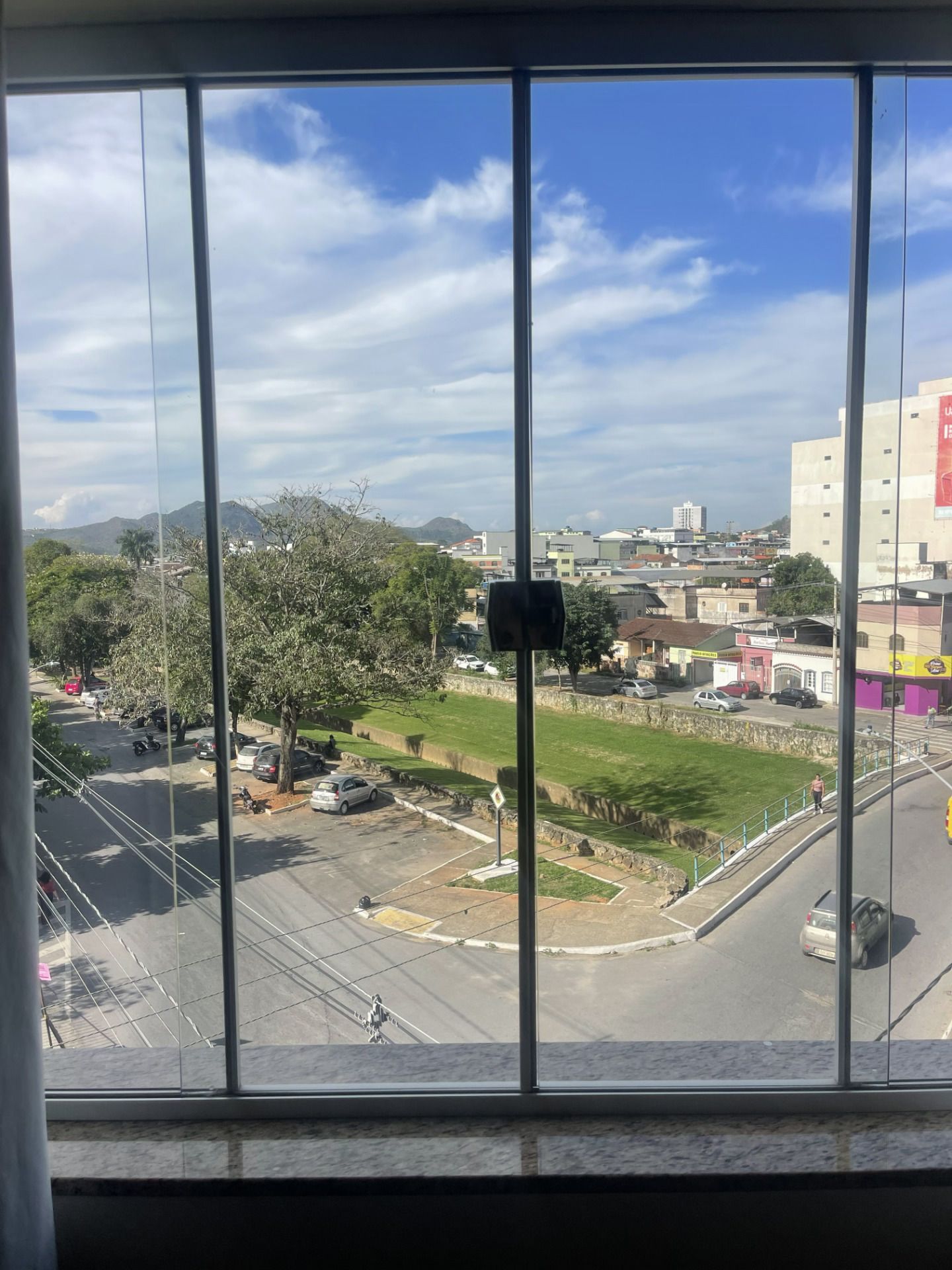 Vista através de uma janela para uma rua da cidade, um terreno gramado, árvores e prédios distantes sob um céu nublado.