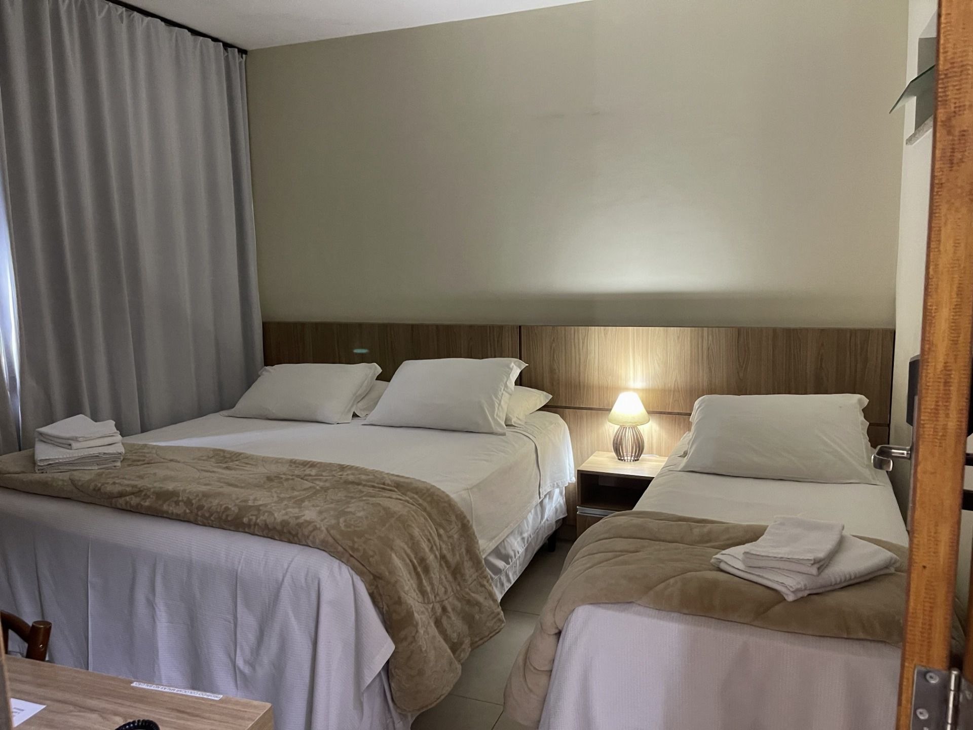 Quarto de hotel com duas camas, lençóis brancos, mantas bege e um abajur aceso perto da janela.