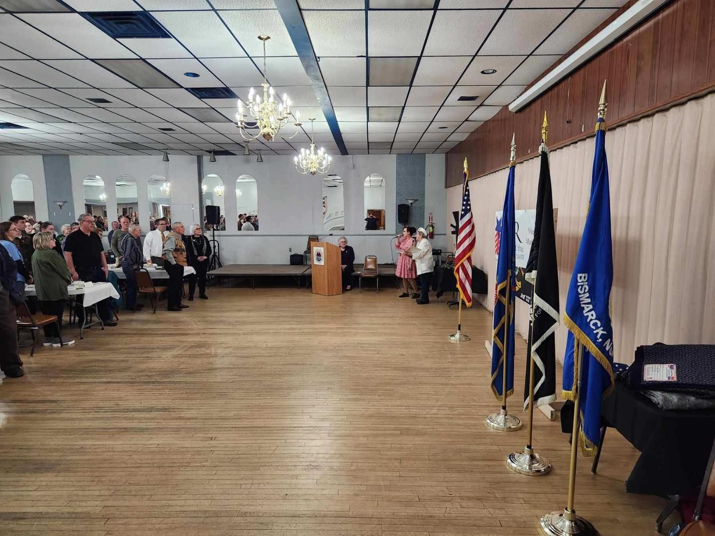 Community hall with seated audience, podium, and flags along the wall on a wood floor