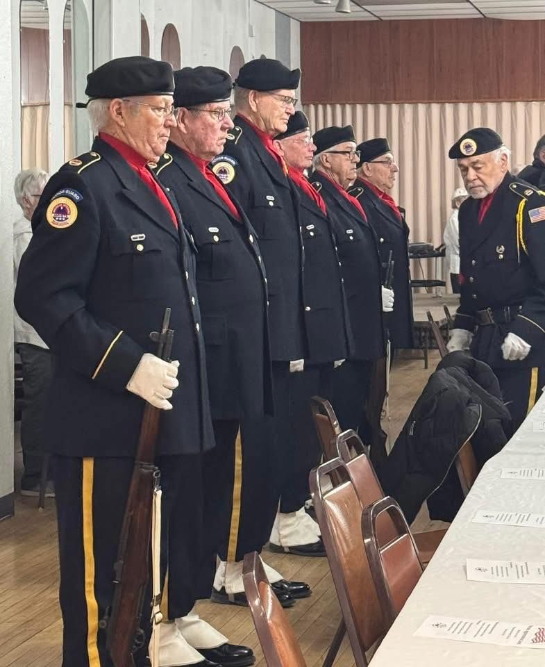 Uniformed veterans standing at attention beside a row of chairs in a hall