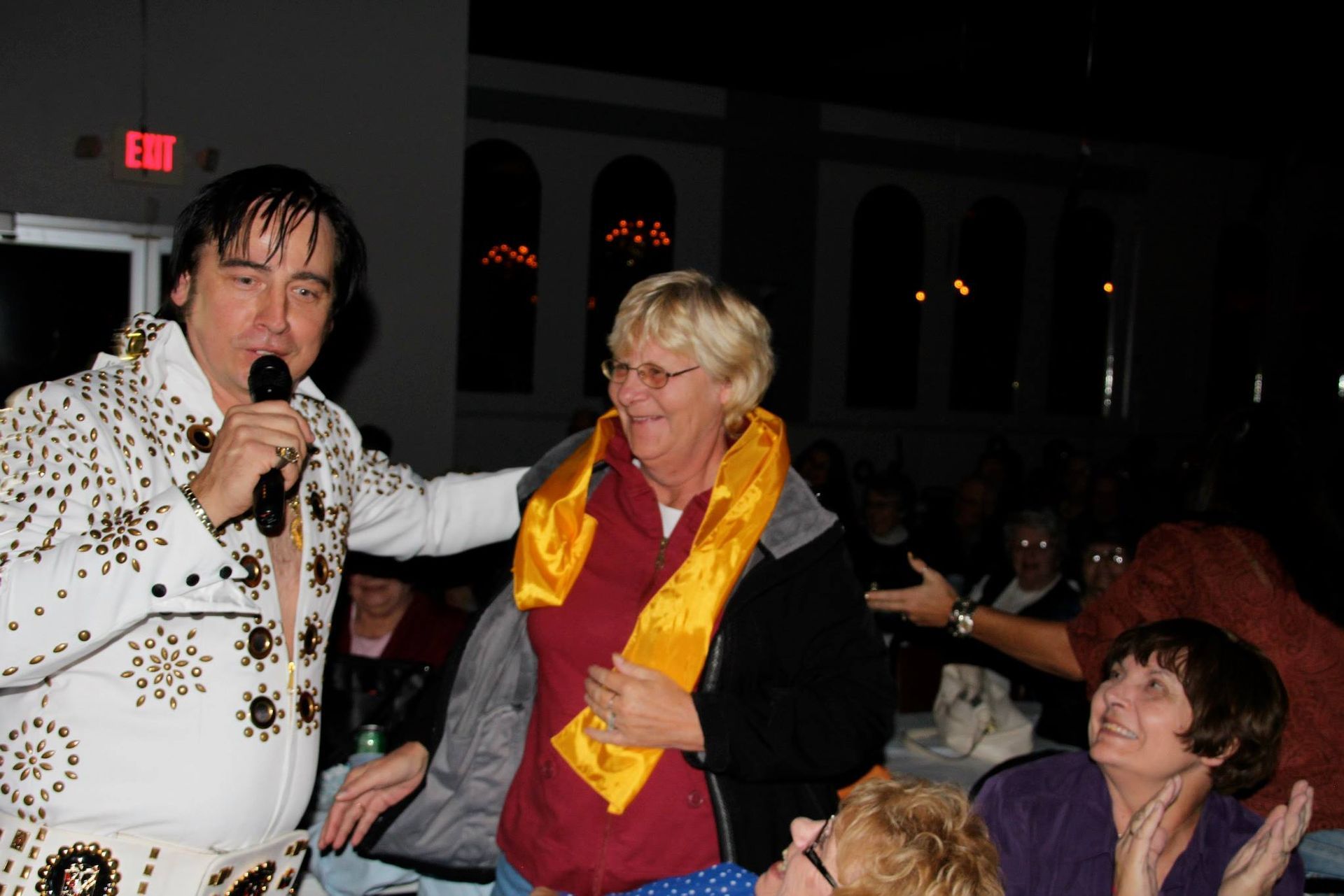 Man holding a microphone beside a smiling person in a yellow scarf at a lively indoor event