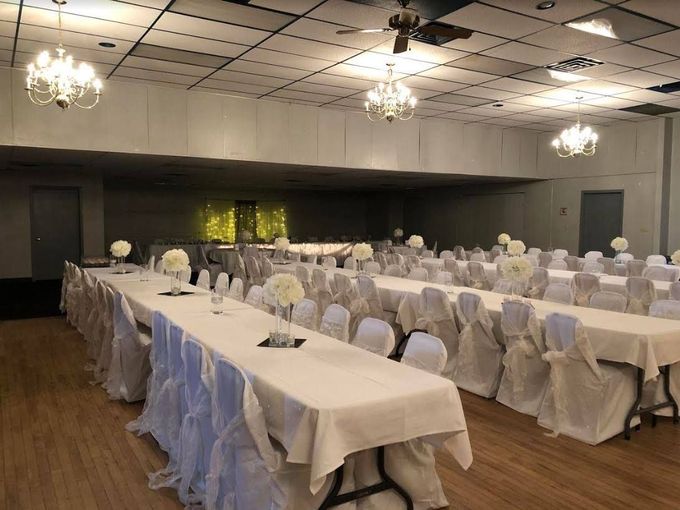 Empty banquet hall with white-covered tables, chairs, and chandeliers under a patterned ceiling