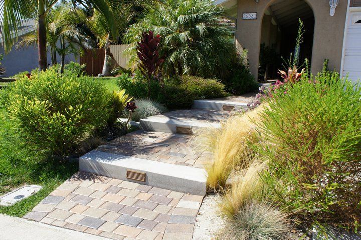 Brick pathway and steps leading to a house entrance, flanked by greenery, shrubs, and plants.