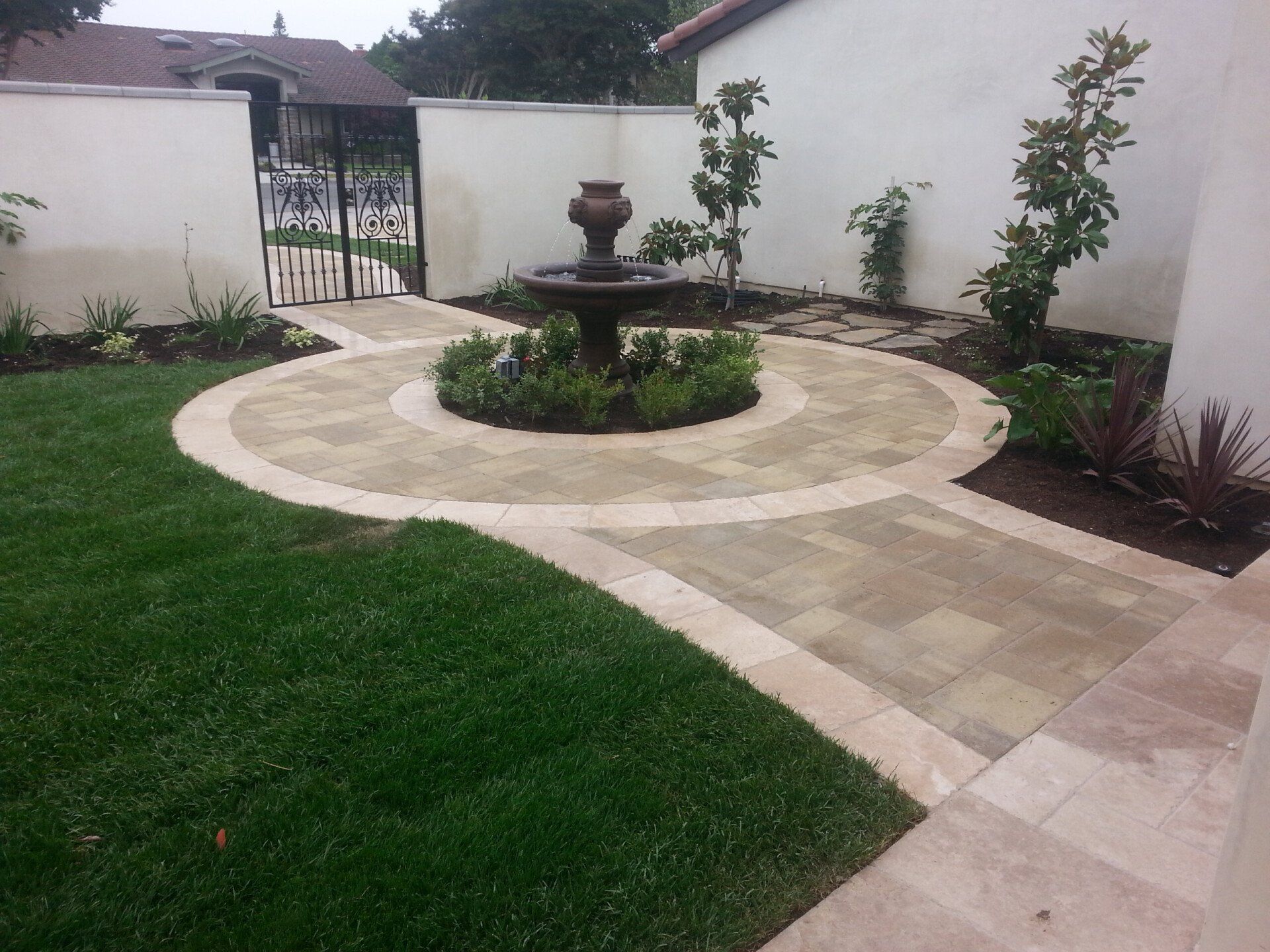 Stone walkway with fountain in a garden, framed by a white wall and green lawn.