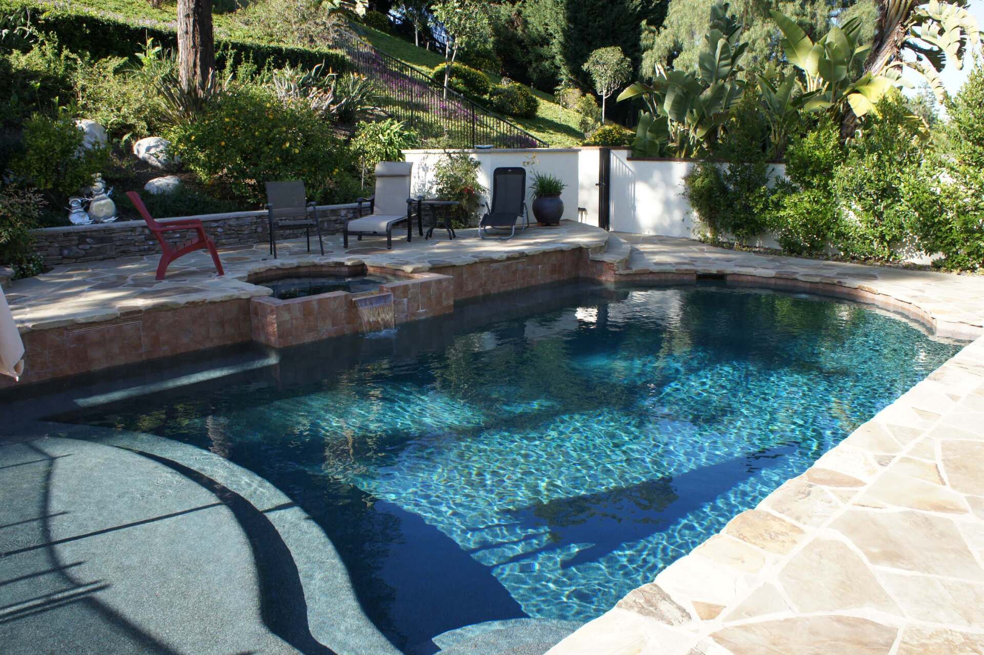 Pool with blue water, stone deck, chairs, and lush greenery in a backyard setting.