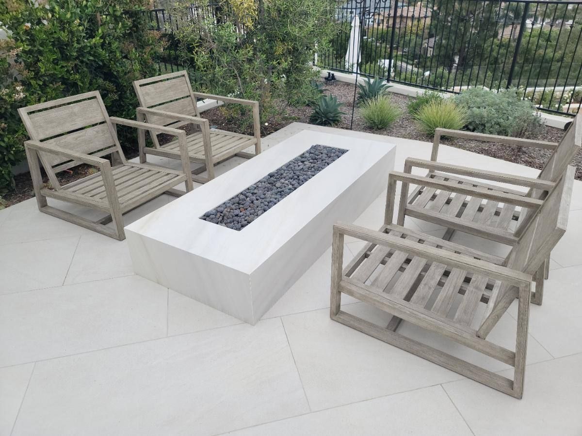 Patio with rectangular fire pit, four armchairs, light-colored pavers, and a view of foliage and fence.