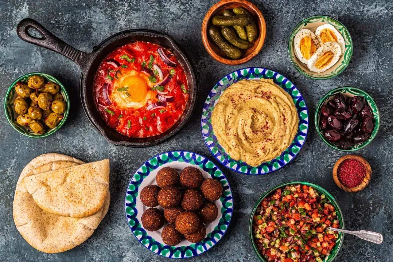 Comida kosher con shakshuka, hummus, falafel, pan pita, huevos, aceitunas, pepinillos y ensalada.