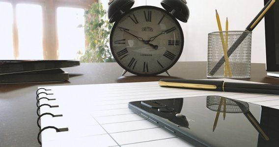 Orologio, penna, telefono e blocco note su una scrivania, con la luce del giorno e il verde visibili attraverso la finestra.