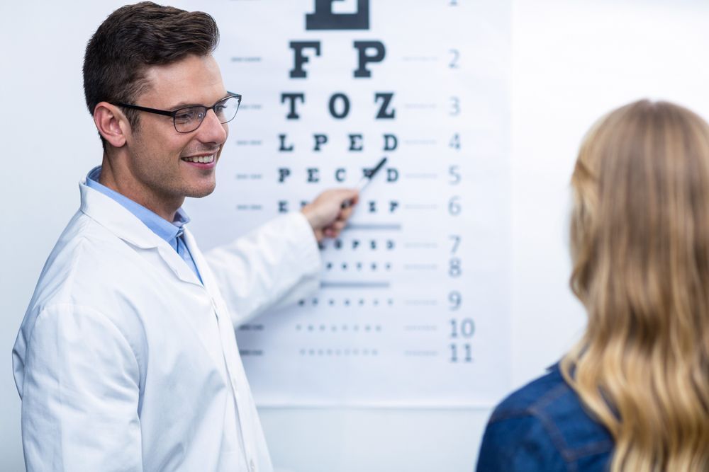 An Optometrist in a White Coat Points to a Snellen Eye Chart — Peninsula Eyecare Izzards Optometrists in Gosford, NSW