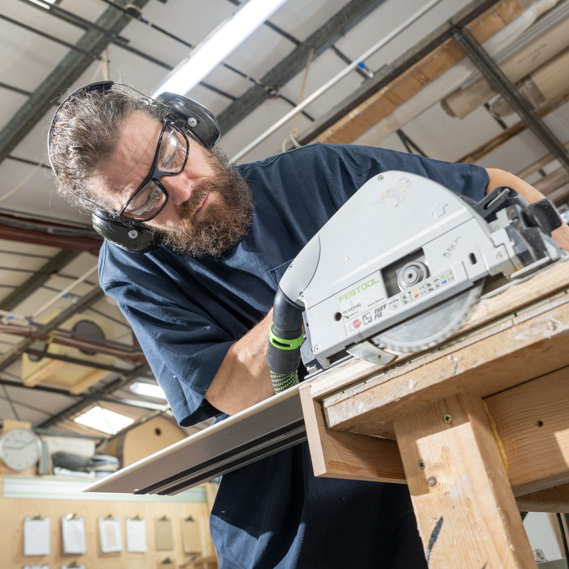 Man using a circular saw to cut wood at a workshop bench, wearing safety glasses and earmuffs