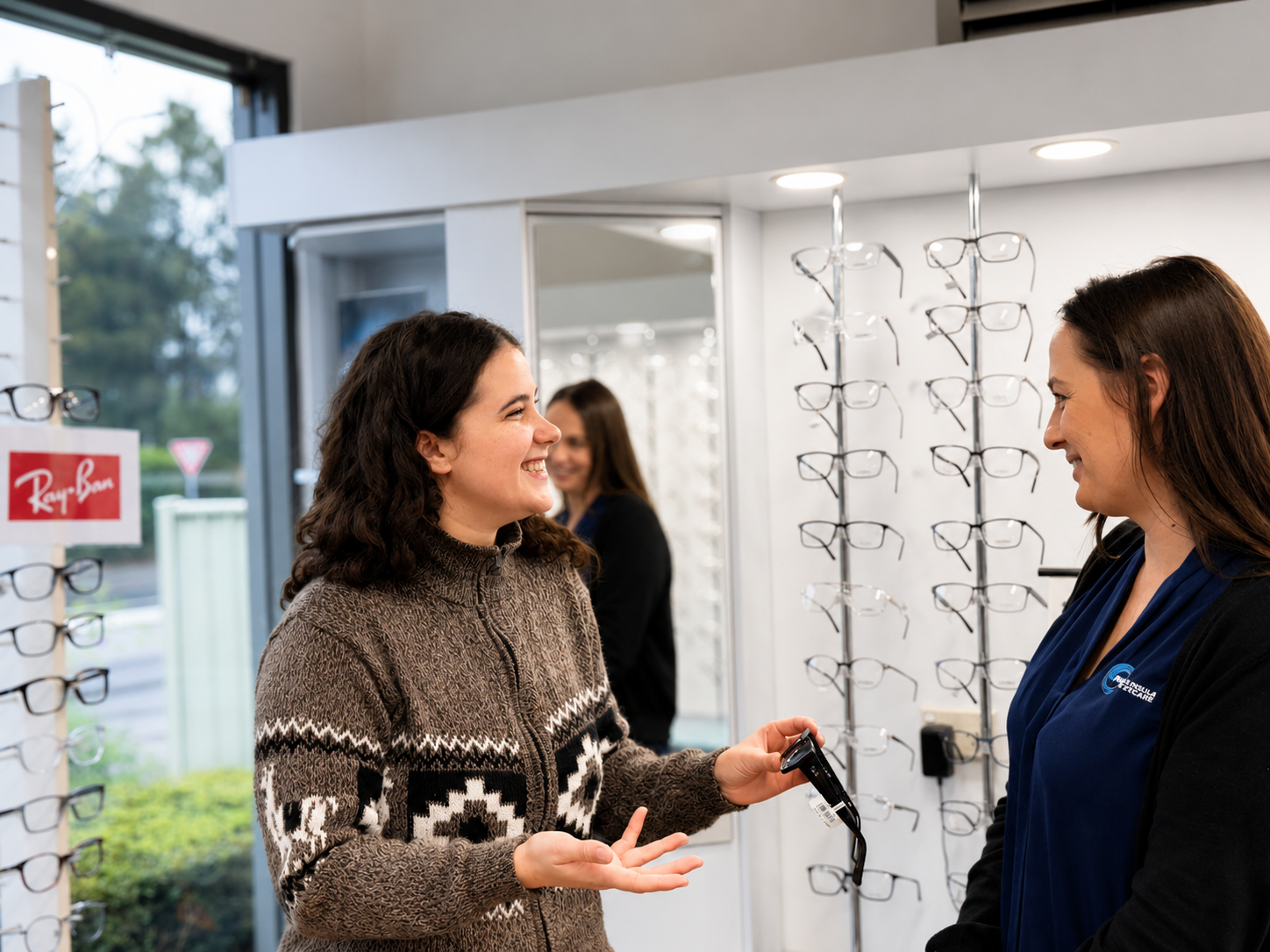 Two women discussing eyeglasses in an optical store, with a display rack of frames nearby.— Peninsula Eyecare Izzards Optometrists in Woy Woy, NSW