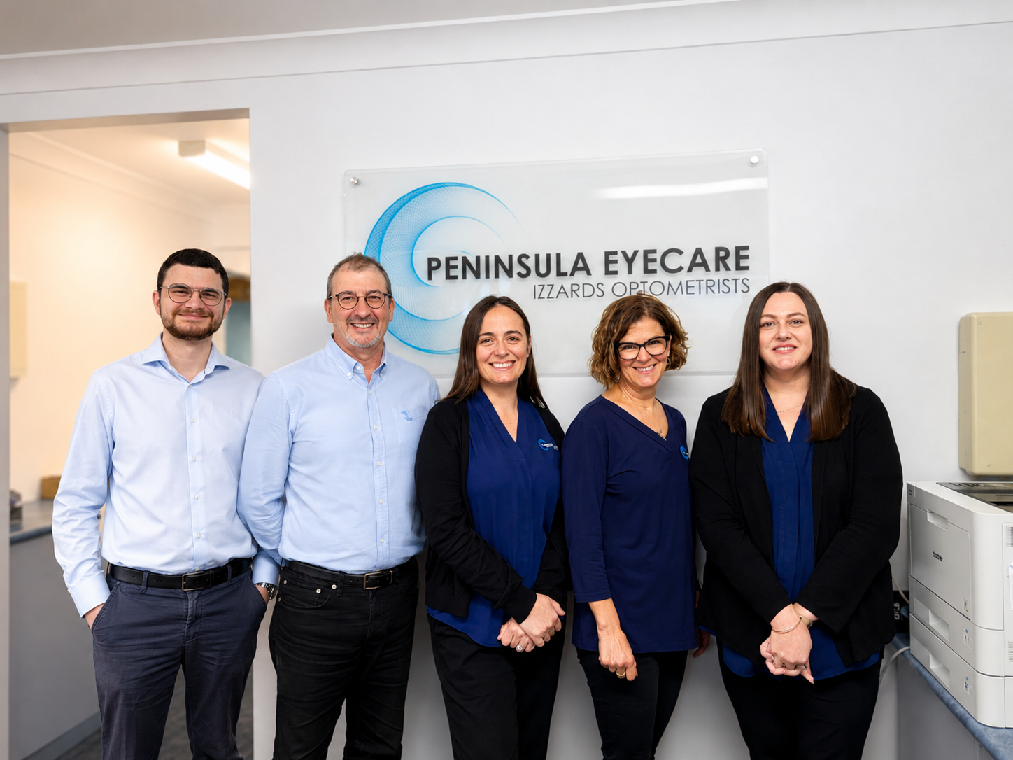 Five people standing in a clinic lobby in front of a Peninsula Eyecare sign— Peninsula Eyecare Izzards Optometrists in Woy Woy, NSW