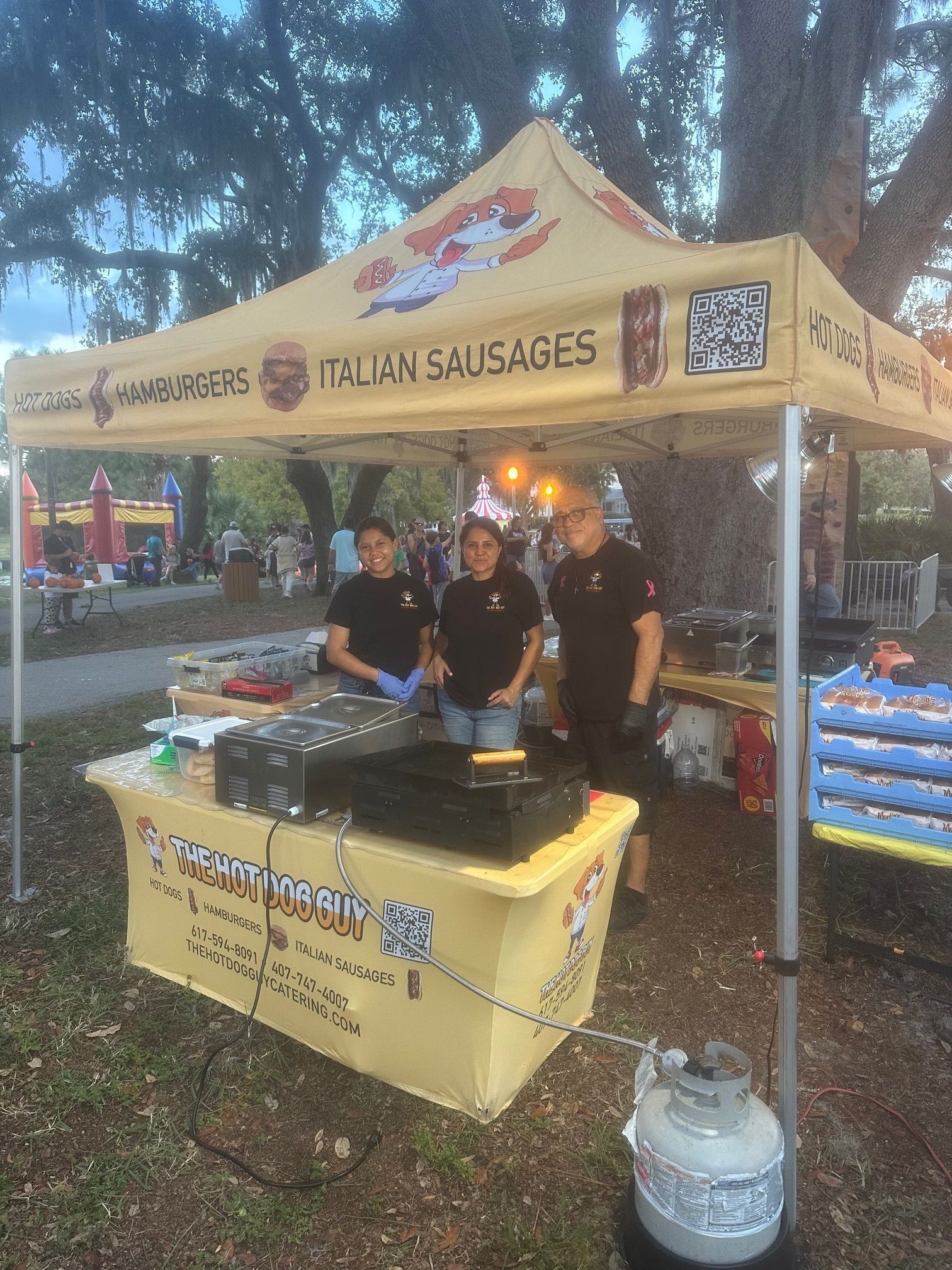 Three food stand workers in black shirts stand behind a table with a grill under a yellow tent at an outdoor event.