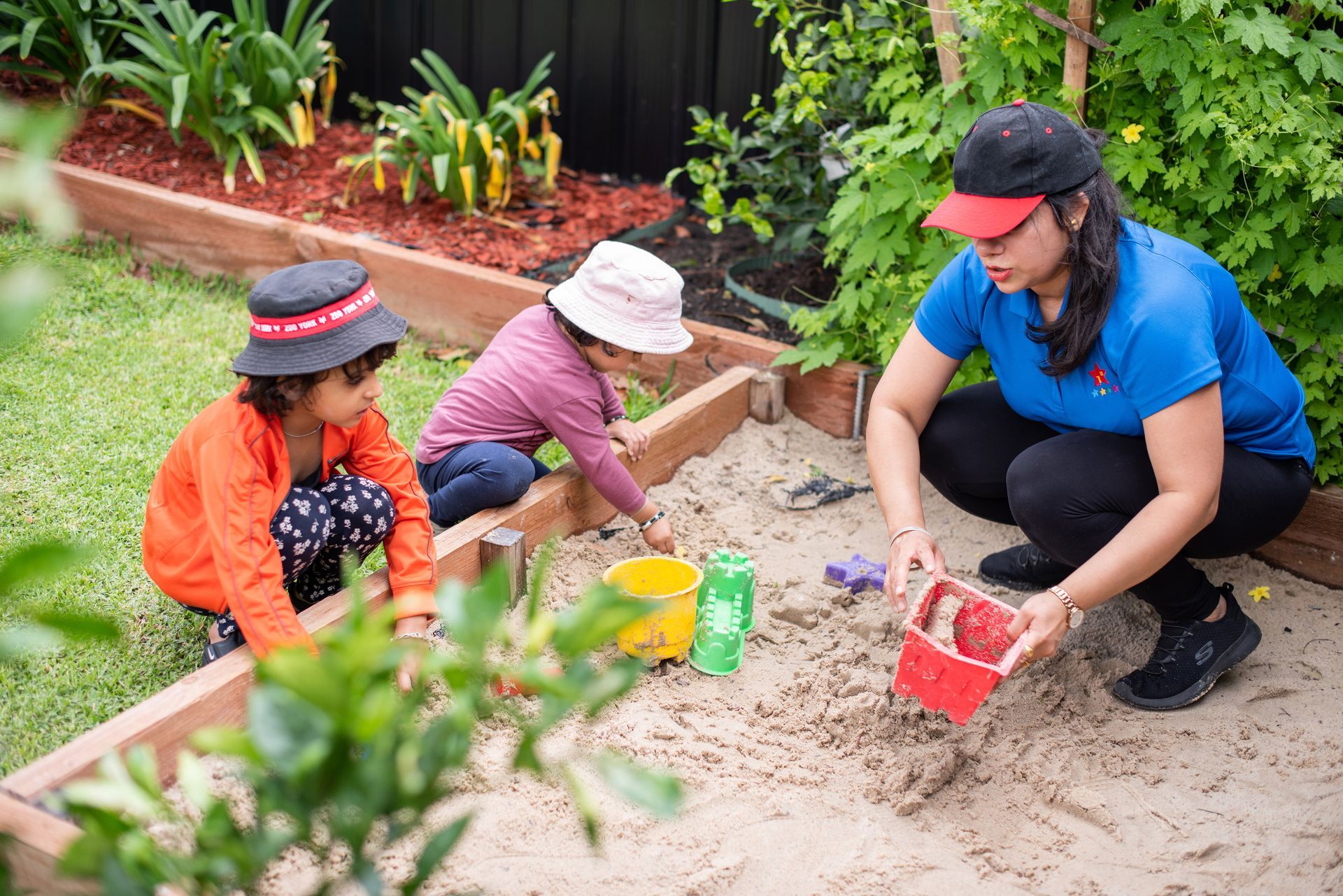 Two children and an adult playing in a sandbox in a garden; using red, yellow, and green buckets.