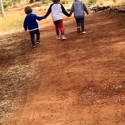 Three children walking hand-in-hand on a red dirt path in a natural outdoor setting.
