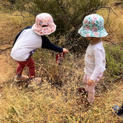 Two young children, wearing sun hats, examine a marker in dry, grassy field.