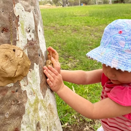 Child in sun hat presses clay onto tree trunk in a park.