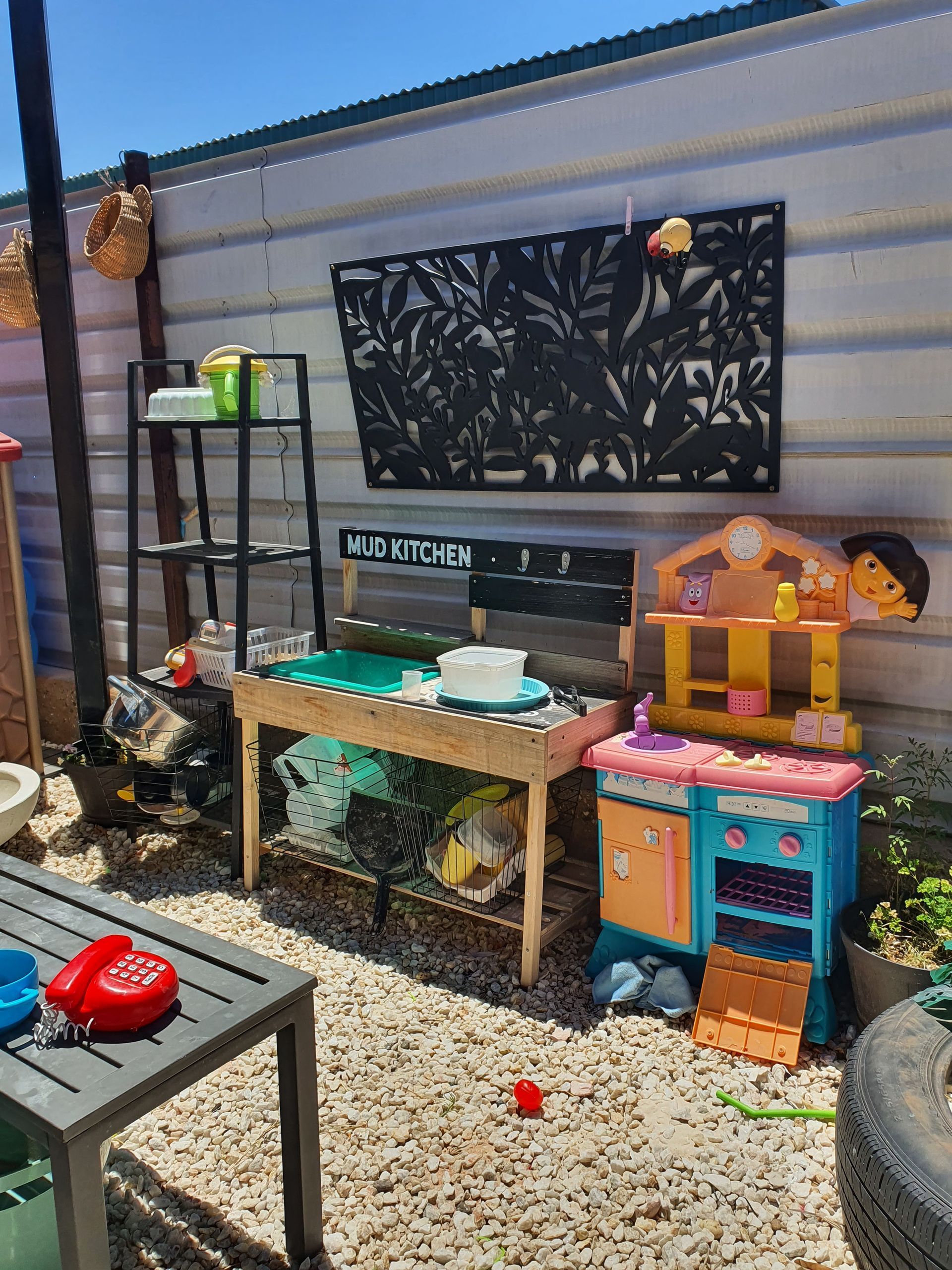 A child's outdoor mud kitchen with toys and a decorative panel on a gravel surface.