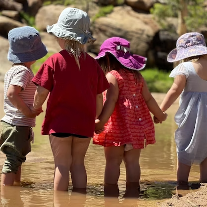 Four young children holding hands, wading in a shallow stream, wearing hats.