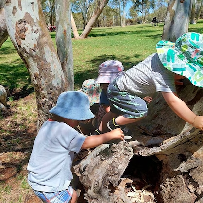 Three children in hats climbing on a fallen tree in a park.