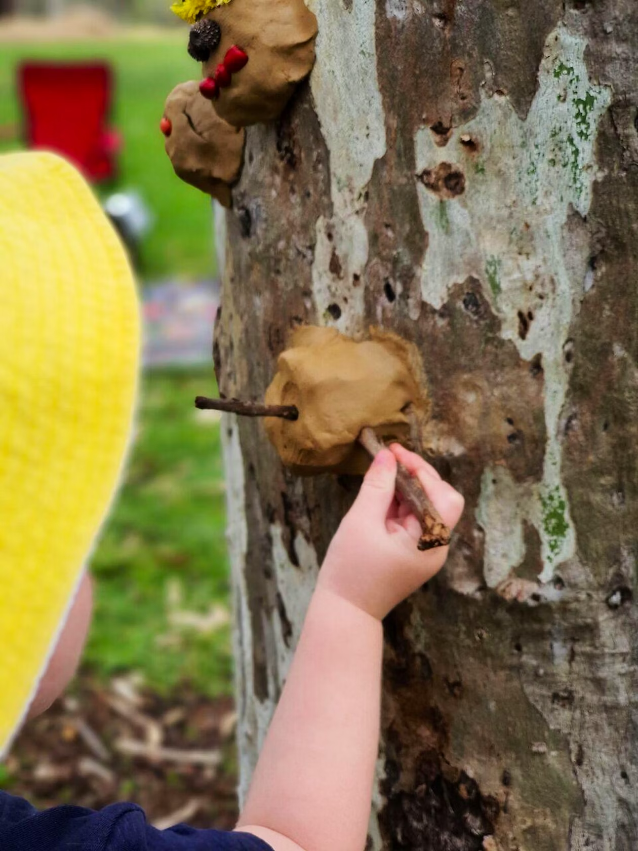Child with yellow hat attaching clay art with stick to a tree trunk outdoors.