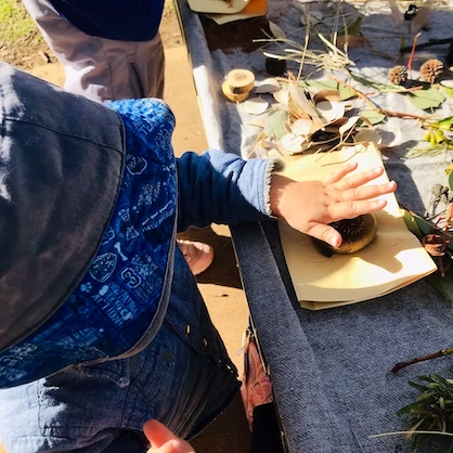 Child pressing a natural object onto paper. Various natural items and craft supplies on a table.