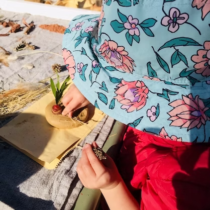 Child wearing a floral hat and red skirt, crafting with a small plant in a sunny outdoor setting.