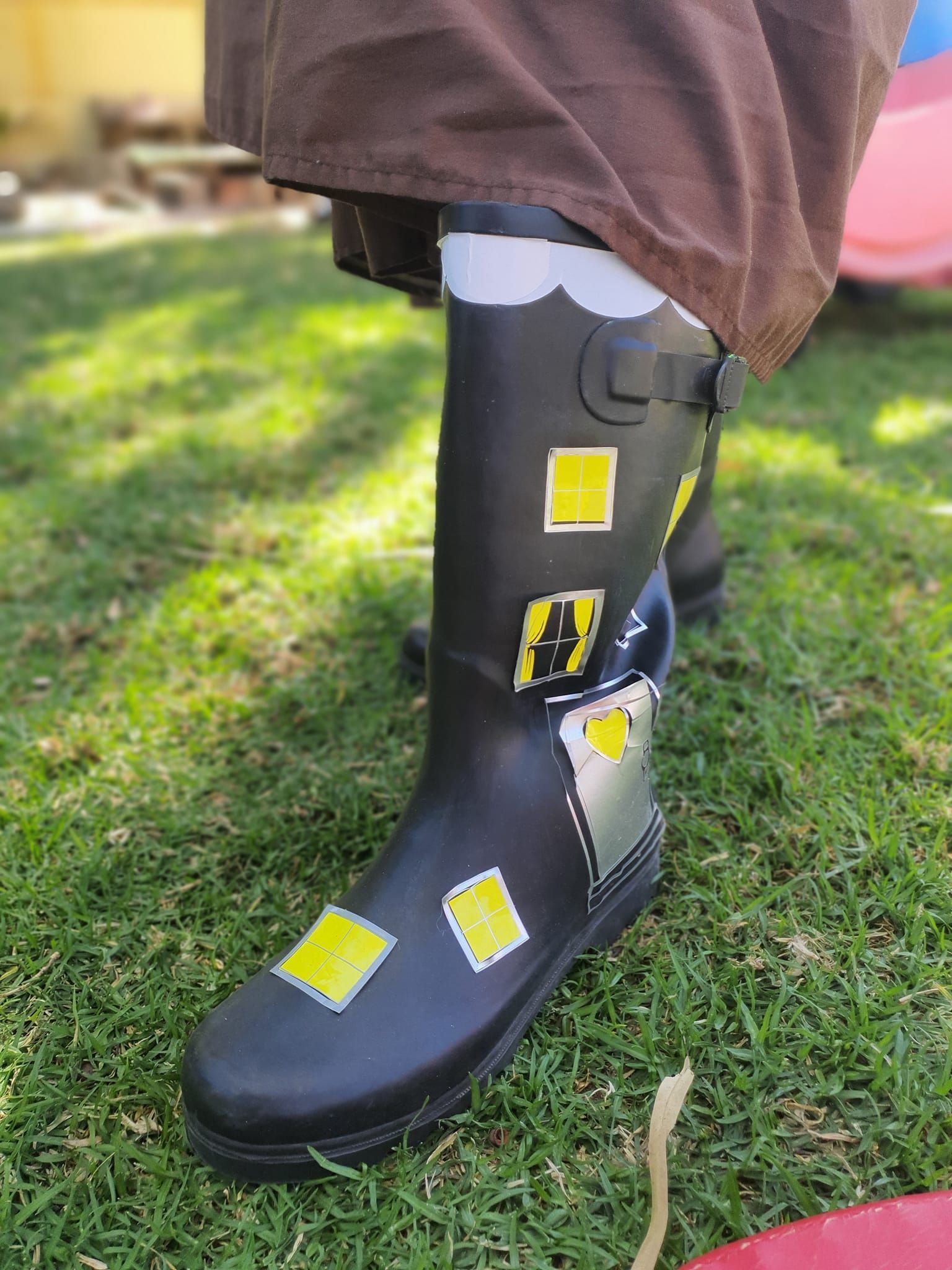 Black rain boot with hand-painted yellow windows and a white cloud design, in a grassy yard.
