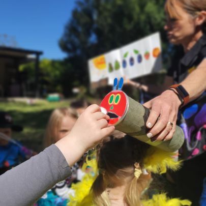 Child feeding a Very Hungry Caterpillar craft while woman reads book outdoors.
