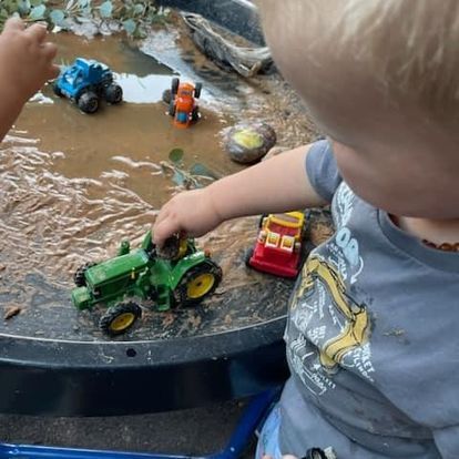 Child playing with toy tractors in a muddy water table, other children present.