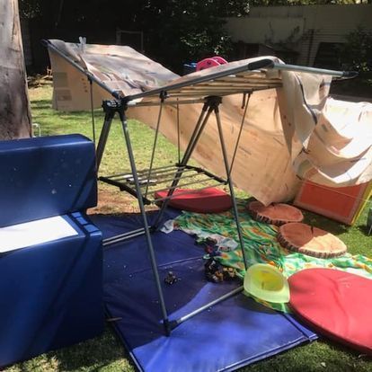 Backyard play area with a metal frame covered with fabric, cushions, and toys on grass.